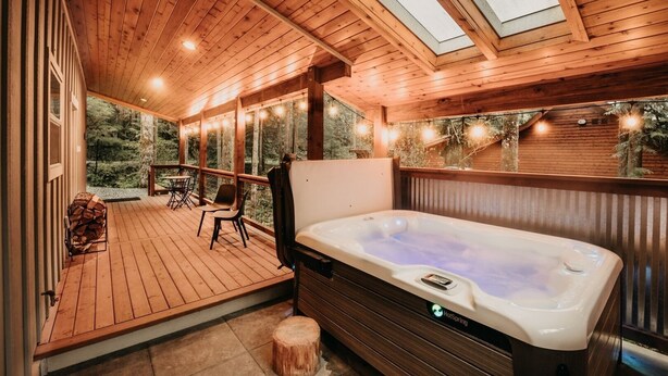 A wooden deck with a hot tub, outdoor seating, and a view of the forest. - 52gs - Hot Tub - Wifi - Pets Ok - Wood Stove - Sleeps 6 2 Bedroom Home by RedAwning (Deming)