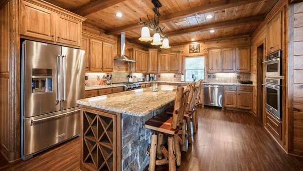 A spacious kitchen with wooden cabinets, a large island with stools, stainless steel appliances, and a stone accent wall. - Breathtaking Views, Seclusion, 4 Bedroom Luxury cabin on 5+ Acres! (Broken Bow)