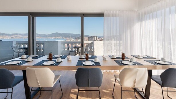 A dining area with a long table set for six, large windows offering a city view, and modern chairs. - Dorma Vigo (Vigo)