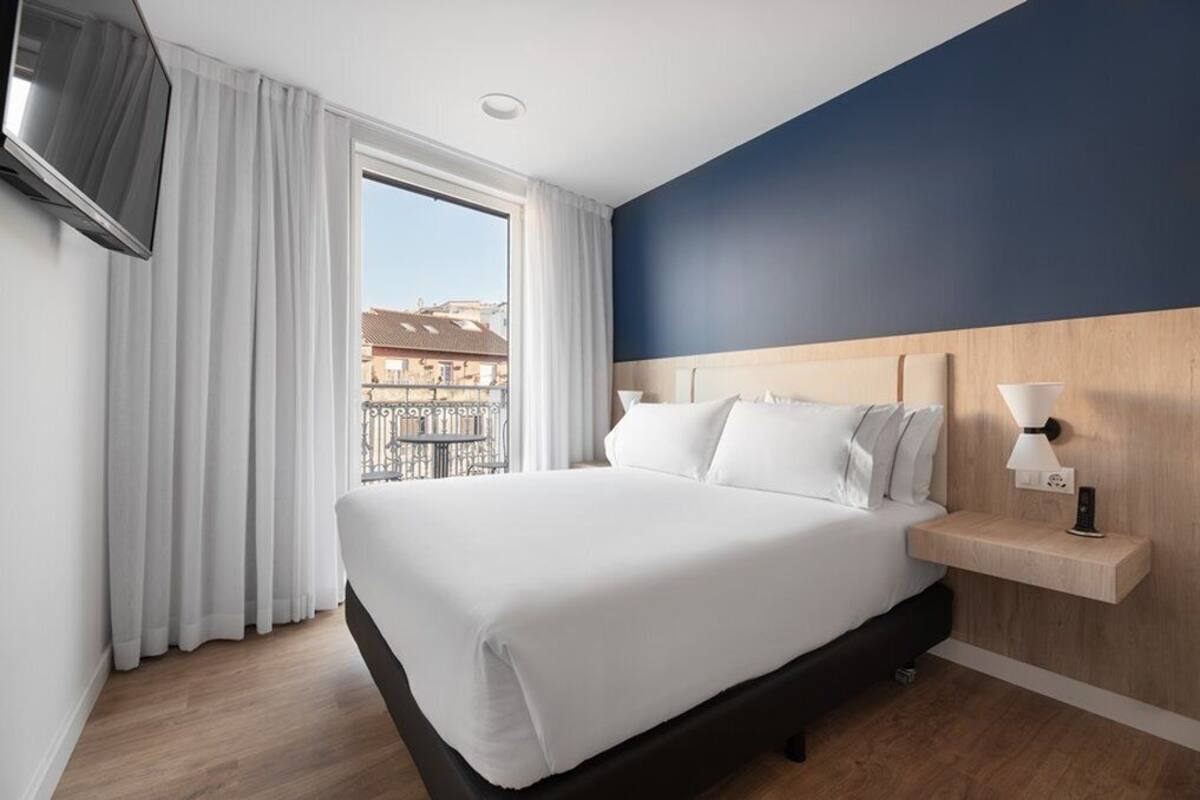 Una habitación de hotel con una cama grande, una mesita de noche de madera, un televisor y una ventana con cortinas.