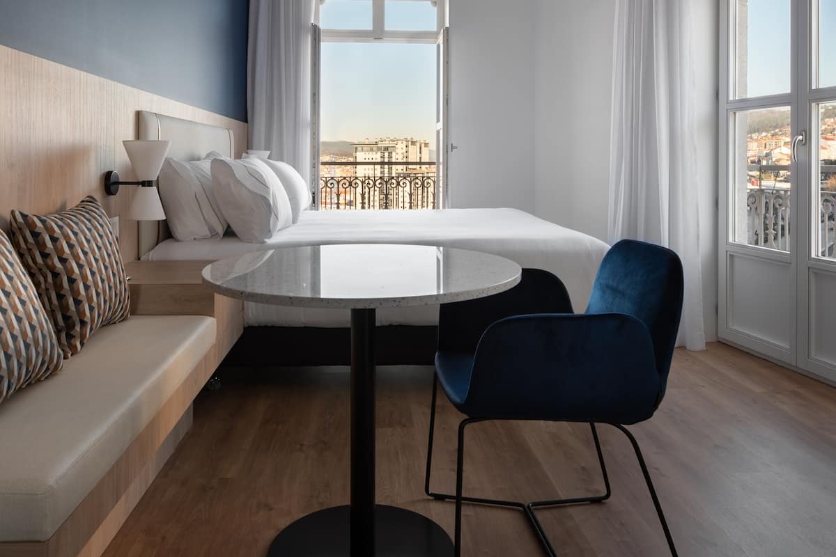 Un hotel moderno con cama, una mesa redonda, una silla azul, un sofá y vistas a la ciudad a través de amplias ventanas.