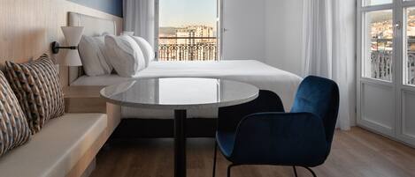 A modern hotel room with a bed, a round table, a blue chair, a sofa, and a view of the city through large windows.