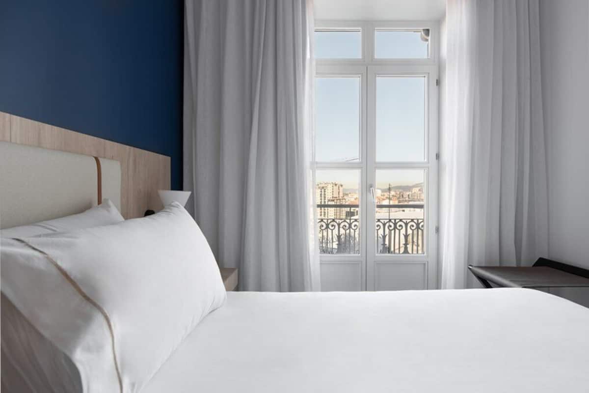 Habitación de hotel con una cama grande, una mesita de noche y vistas de un paisaje urbano a través de una ventana con cortinas.