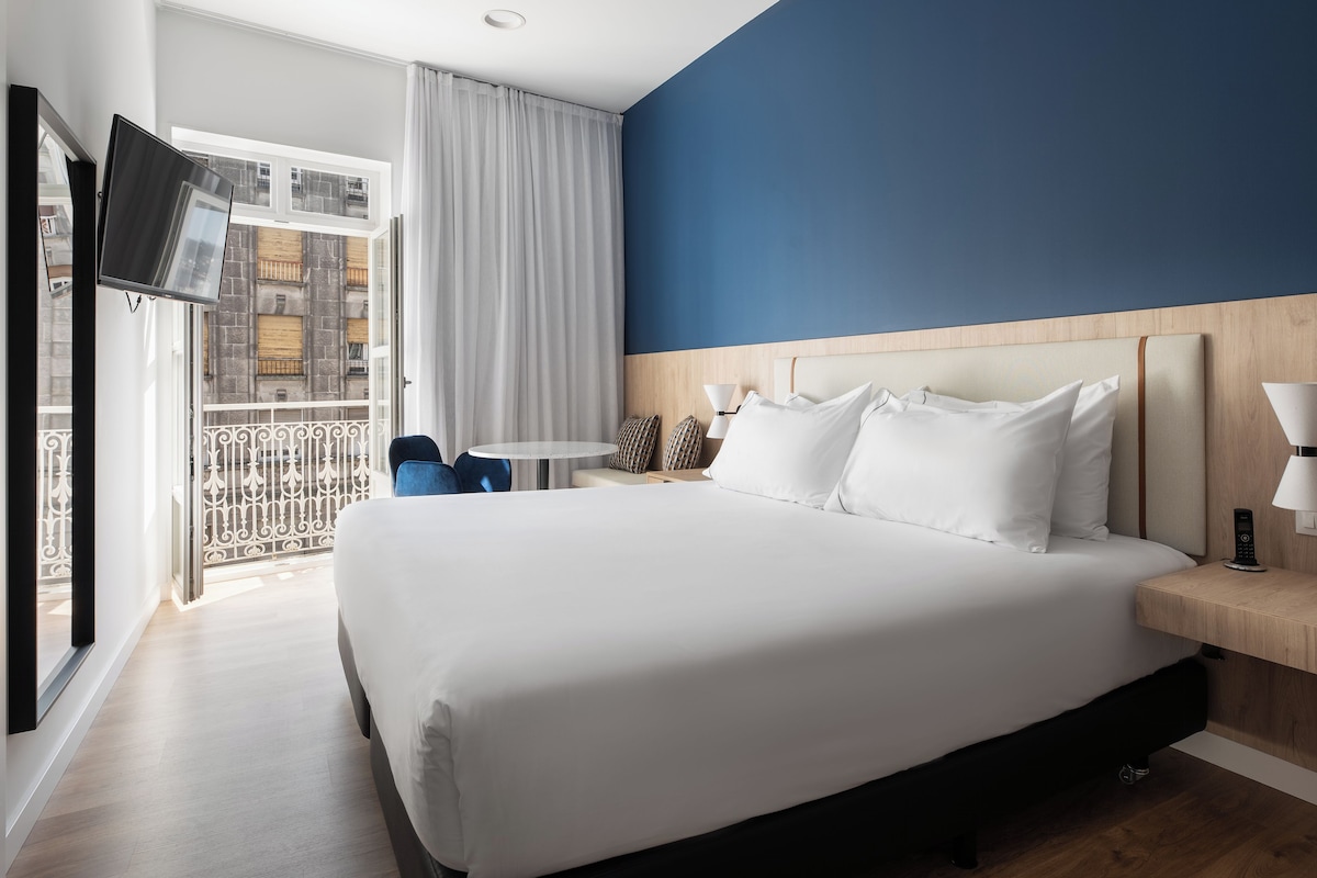 A hotel room with a large bed, a desk, a chair, a TV, and a balcony with a view of buildings.