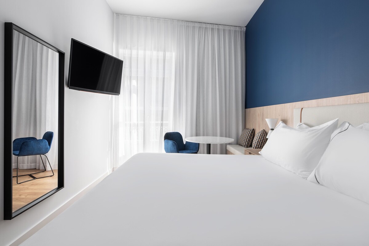A hotel room with a large bed, a TV mounted on the wall, a small round table, two blue chairs, and a wooden headboard.