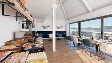 A modern dining area with a buffet setup, a view of the sea, and a high ceiling with exposed beams.