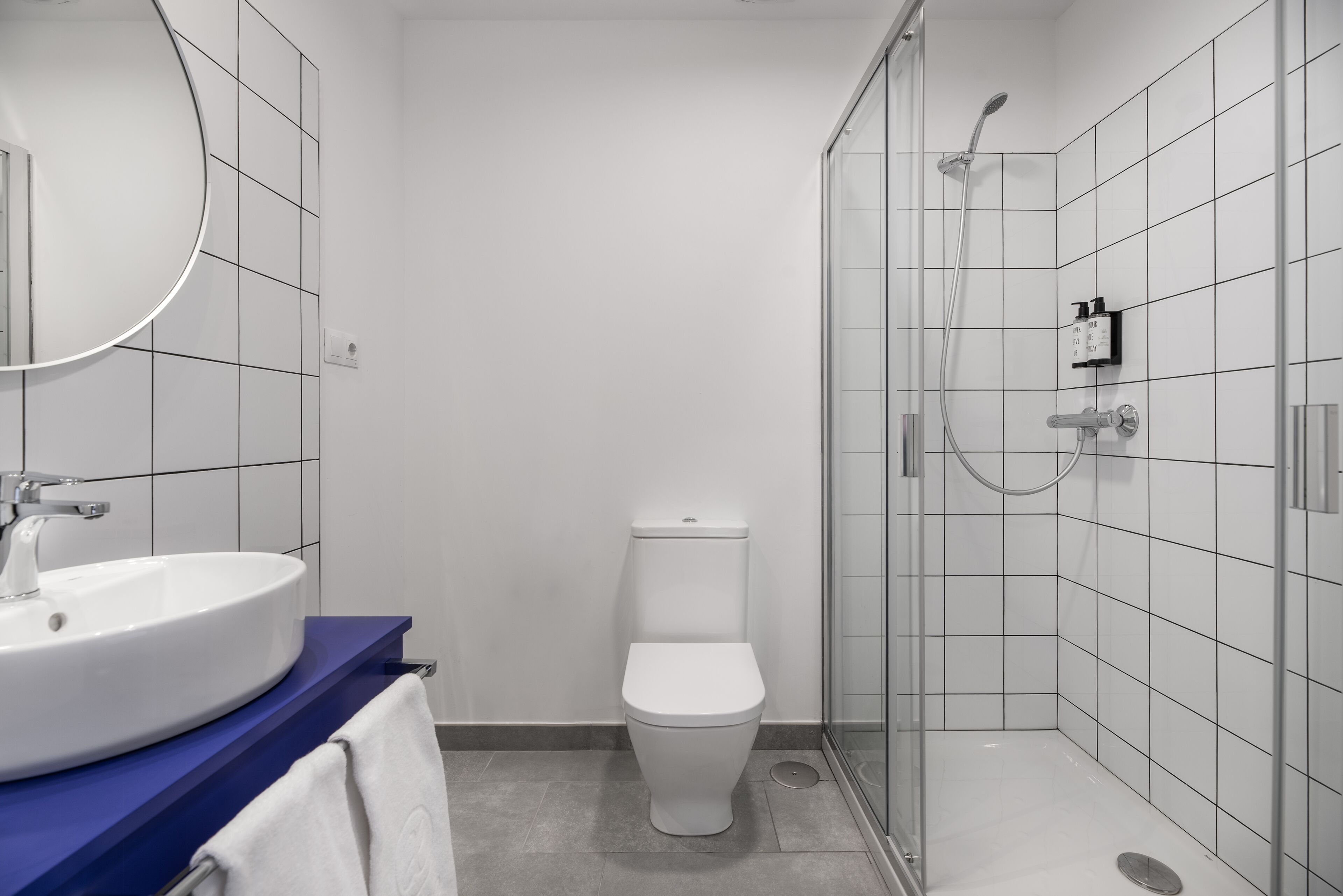 Een moderne badkamer met een wit toilet, een ronde spiegel, een wastafel en een douche met glazen wanden.