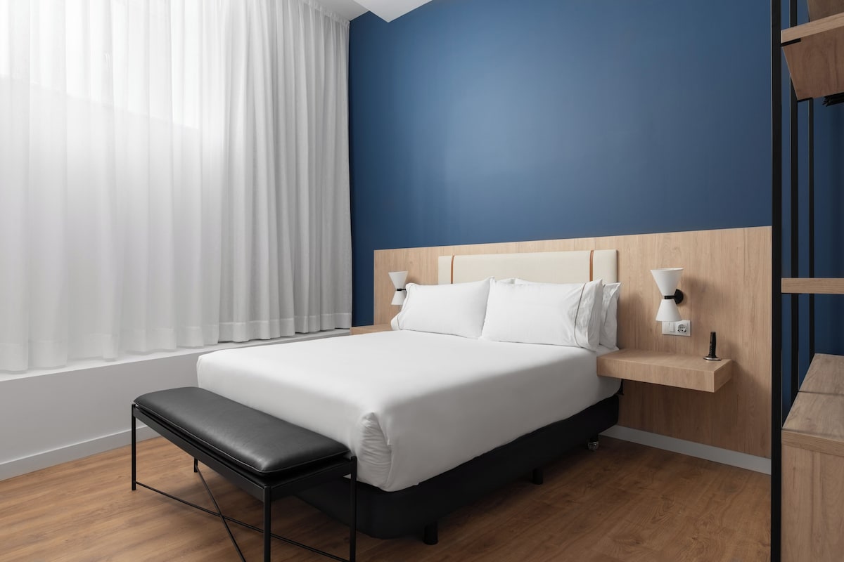A modern hotel room with a large bed, a bench, a bedside table with a lamp, and a view of the window with sheer curtains.