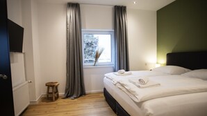 Quarto com cama, janela com cortinas, um banquinho pequeno e uma TV presa à parede.