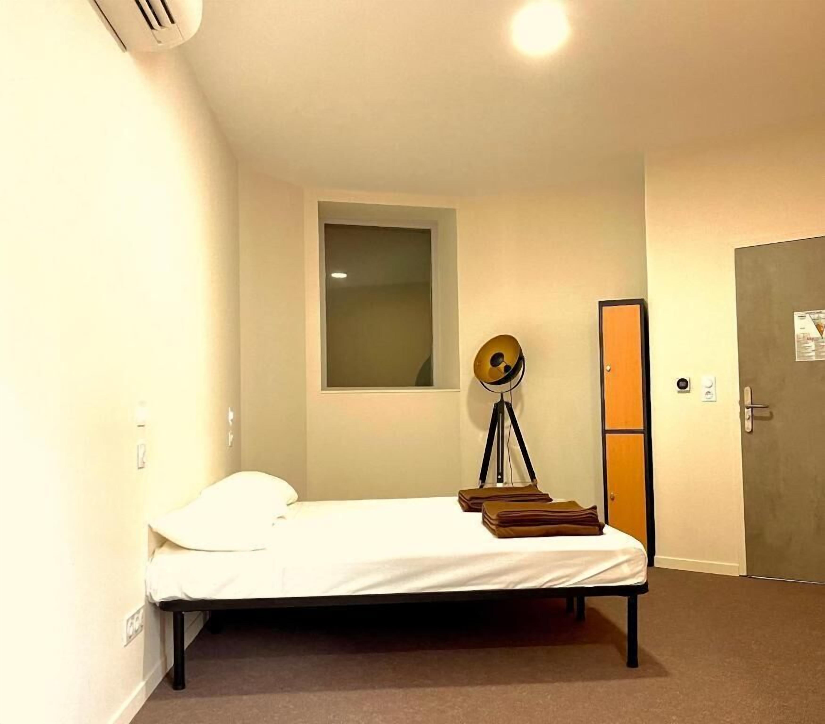 Ein kleines Zimmer mit einem Einzelbett, einem Fenster, einer Stehlampe und einer Tür.
