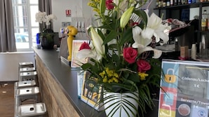 A hotel reception area with a wooden counter, metal stools, a flower arrangement, and a menu board. - Hostel Flamingo Nîmes Centre Gare (Nîmes)