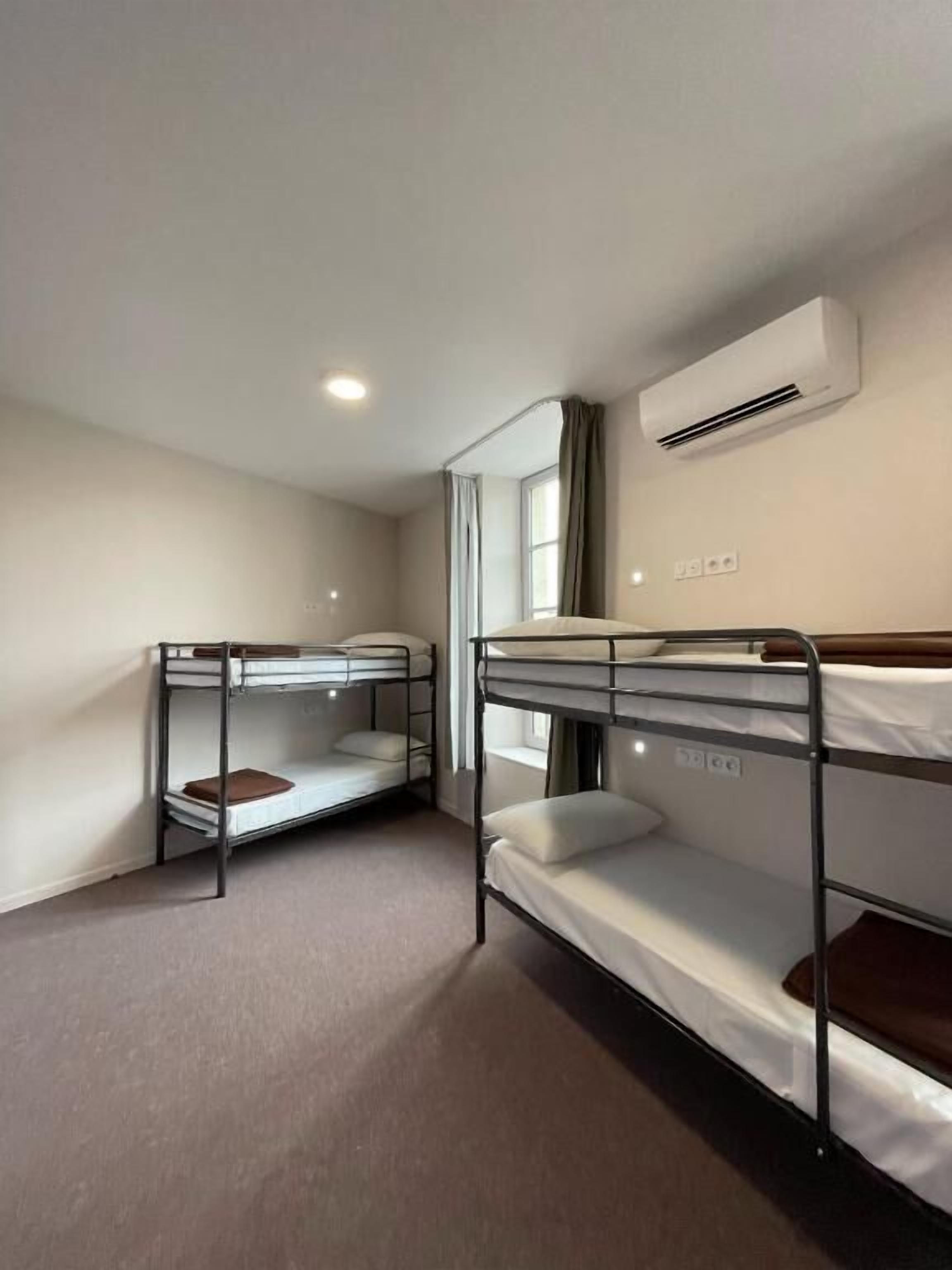 A room with two bunk beds, a window with curtains, an air conditioning unit, and a light switch on the wall.