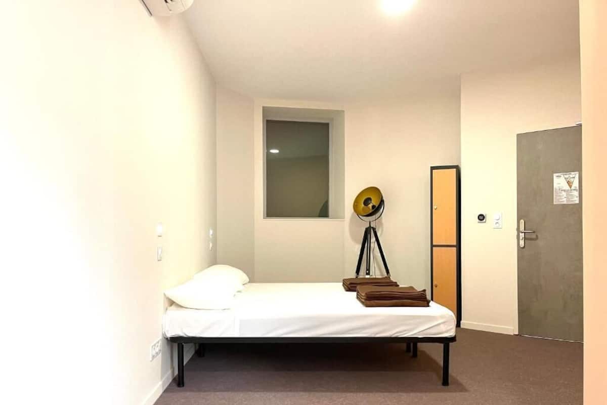 A small, neatly arranged room with a single bed, a window, a door, and a wall-mounted lamp.