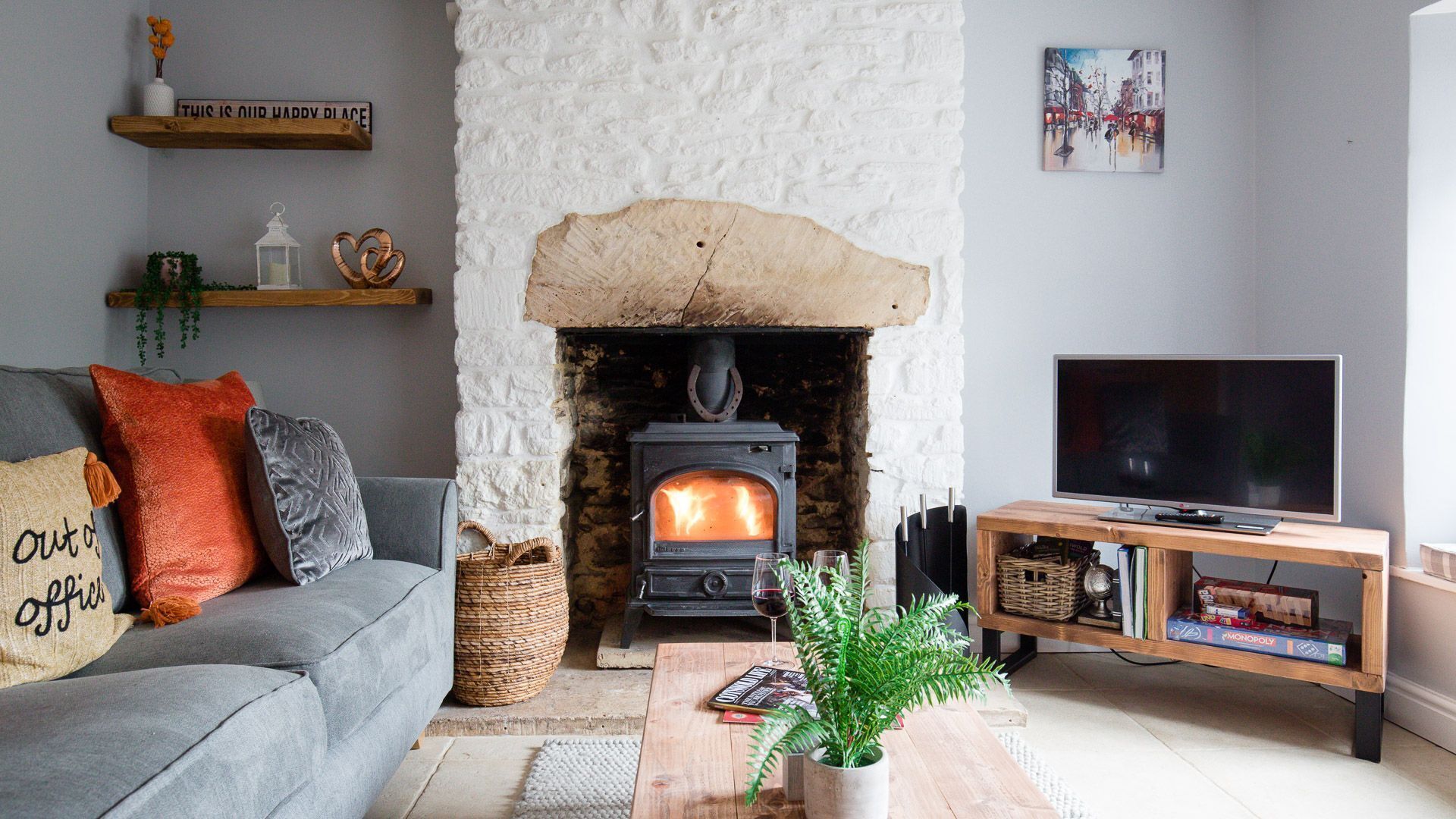 Ein gemütliches Wohnzimmer mit Kamin aus Stein, einem grauen Sofa mit orangefarbenen und grauen Kissen, einem Fernseher auf einem Holzständer und einem Bücherregal mit Gesellschaftsspielen.