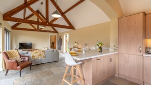 A spacious living area with a high ceiling and exposed wooden beams, a cozy seating area with a leather armchair and a sofa, and a kitchen with wooden cabinets and a bar area.