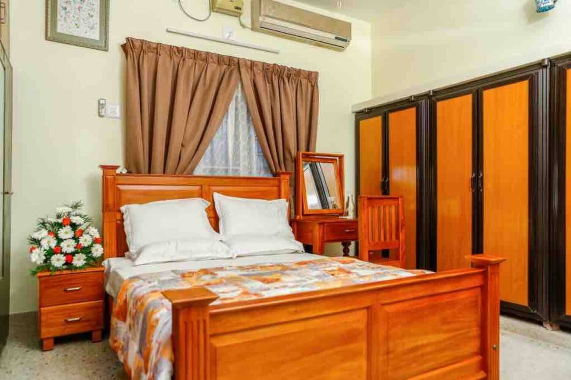 A bedroom with a wooden bed, a nightstand with a floral arrangement, a wardrobe, and a mirror on the wall.