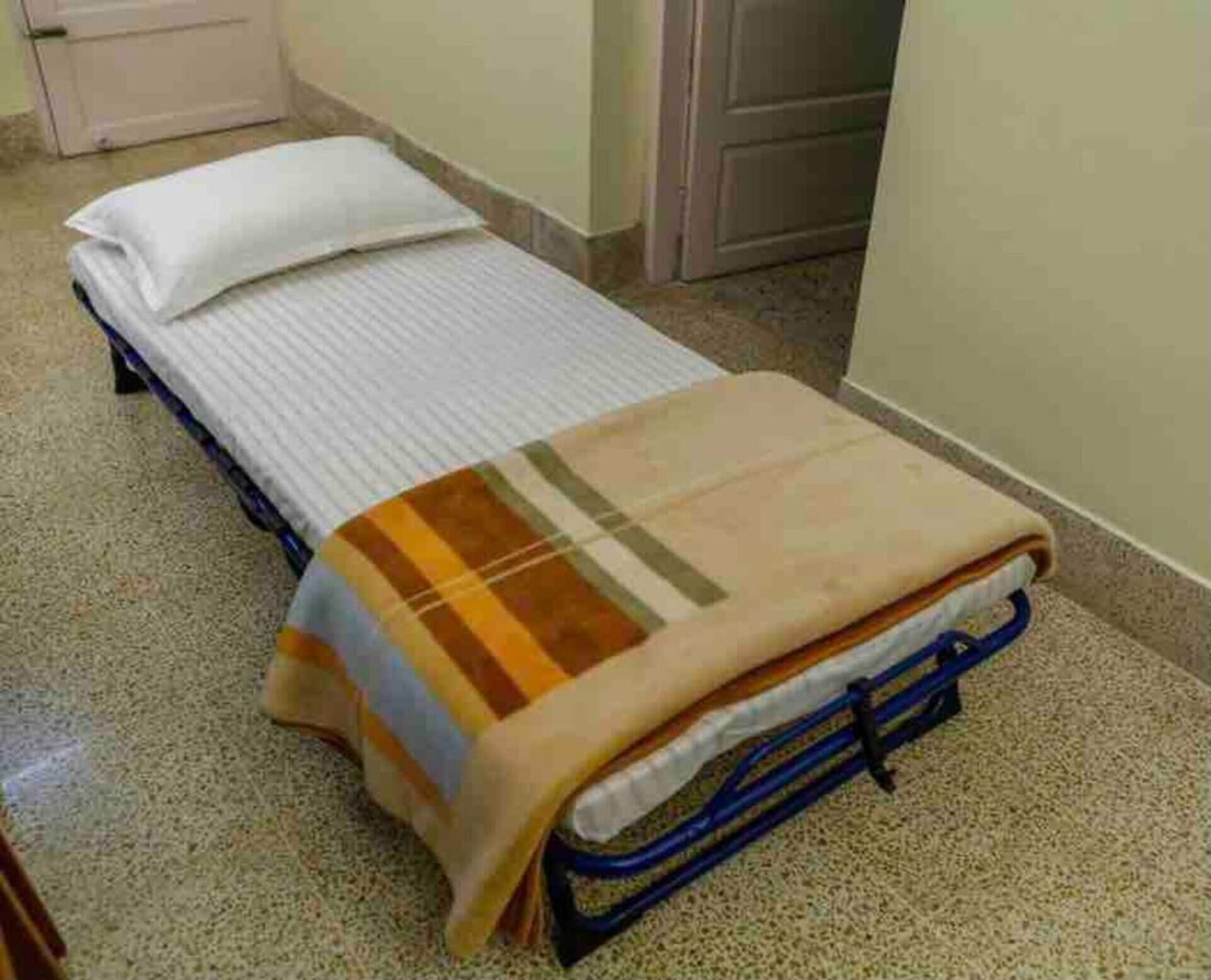 A single bed with a striped blanket and a pillow in a room with a tiled floor and a door in the background.