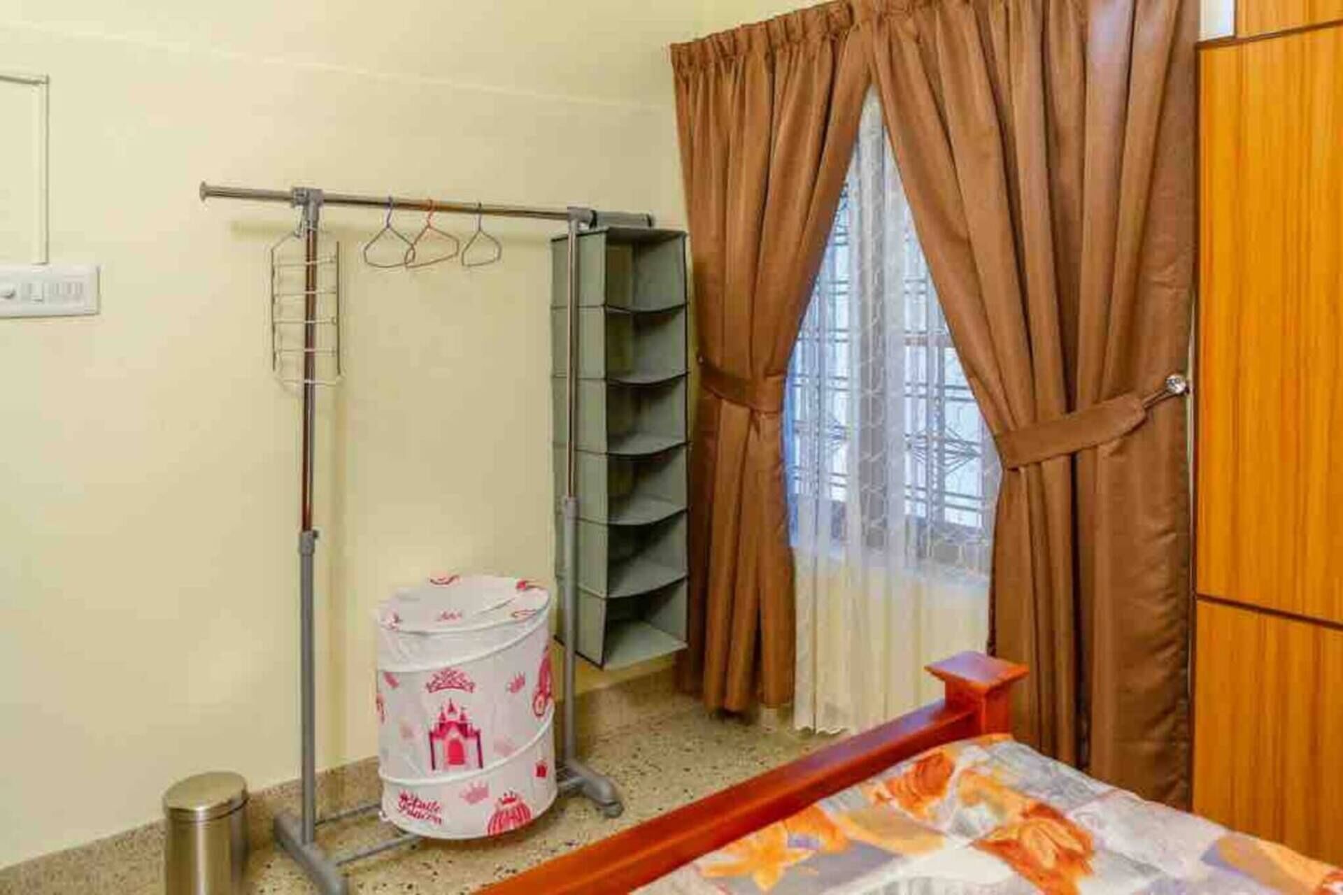 A hotel room with a bed, a metal rack with hangers, a window with curtains, and a trash bin.