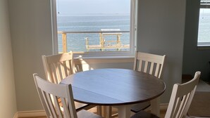 A dining area with a round table and white chairs, a window with a view of the ocean, and a ceiling light fixture.