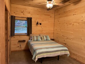 A cozy wooden cabin bedroom with a bed, a window with curtains, a ceiling fan, and a wall-mounted lamp. - Hocking Hills (Buck Crossing Retreat) (Rockbridge)