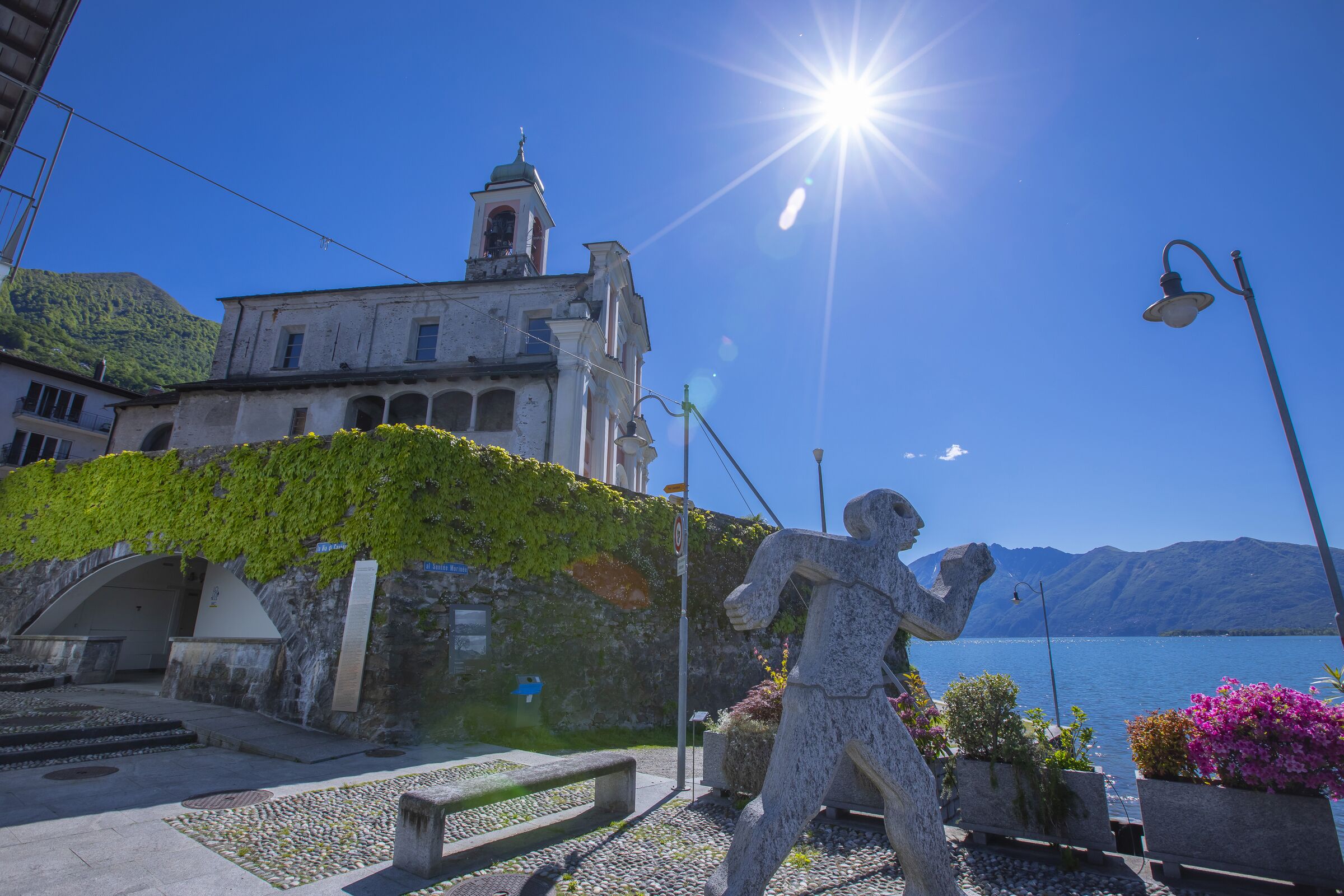 Eine Steinstatue auf einem öffentlichen Platz, im Hintergrund eine Kirche und Berge.