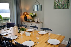 A dining area with a table set for six, chairs, a lamp, a vase with flowers, and a view of the outdoors.