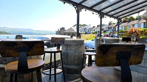 A covered outdoor seating area with wooden chairs and tables, overlooking a body of water and residential buildings.