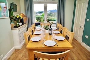 A dining room with a wooden table set for six, chairs, a vase with flowers, and a view through the window.