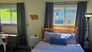 A bedroom with a bed, a desk, and a chair. There is a window with curtains, a lamp, and a map on the wall.