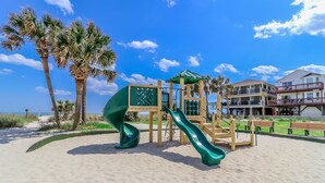 A beach playground with a slide and climbing structures, surrounded by palm trees and a building with balconies. - Ocean Lakes site 6013 beautiful 3 bedroom quiet street 2 carts available (Myrtle Beach)