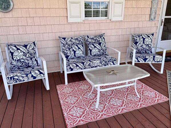 A patio with white chairs and a table, a red patterned rug, and a clock on the wall.