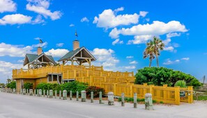 A yellow building with a wooden fence, a palm tree, and a clear blue sky. - Ocean Lakes site 6013 beautiful 3 bedroom quiet street 2 carts available (Myrtle Beach)