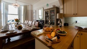 Uma área de refeições com uma longa mesa de madeira posta para uma refeição, um balcão de cozinha com comida e bebidas, e um armário com portas de vidro.