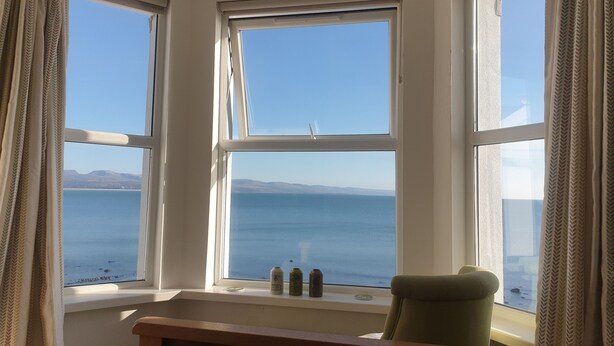 Quarto com janela saliente com vista para um corpo d'água e terra distante. Há uma cama com colcha florida, uma cadeira e duas latas no parapeito da janela.