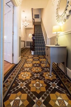 Um corredor com piso estampado, escada, porta branca, luminária e espelho na parede.