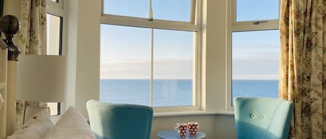 A room with a bed, two blue armchairs, a small table with two mugs, and a view of the sea through large windows.