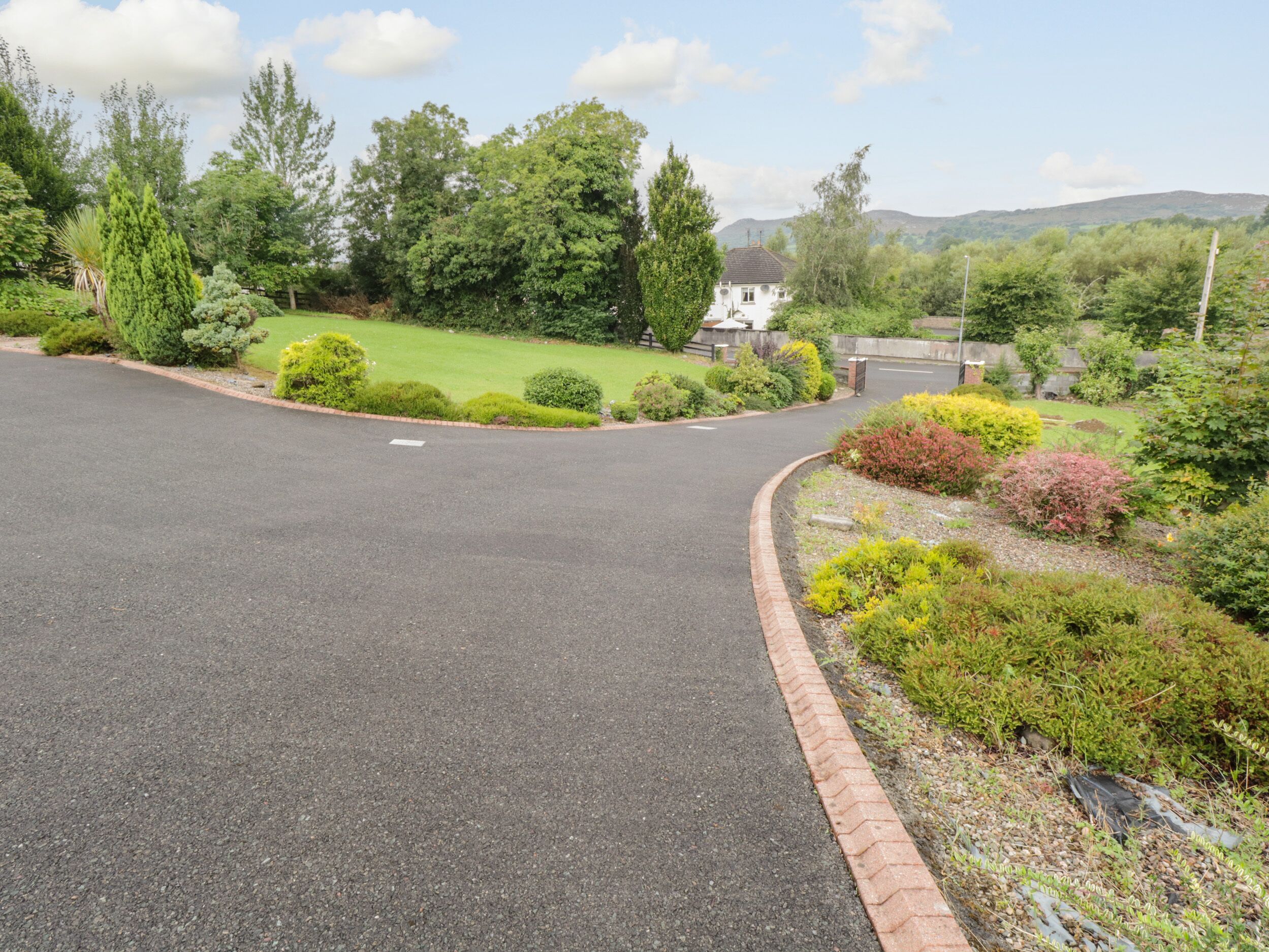A residential street with a paved road, a brick curb, and a variety of well-maintained gardens and trees.