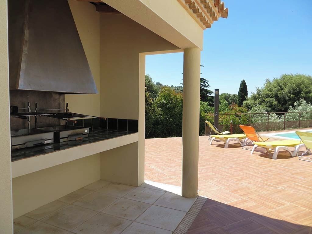 An outdoor kitchen area with a chimney, a poolside lounge with sun loungers, and a view of the surrounding greenery.