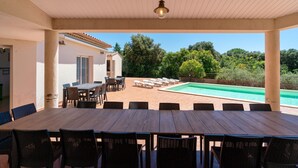 Un patio exterior con mesa y sillas de madera, una piscina y vistas a árboles y una casa.