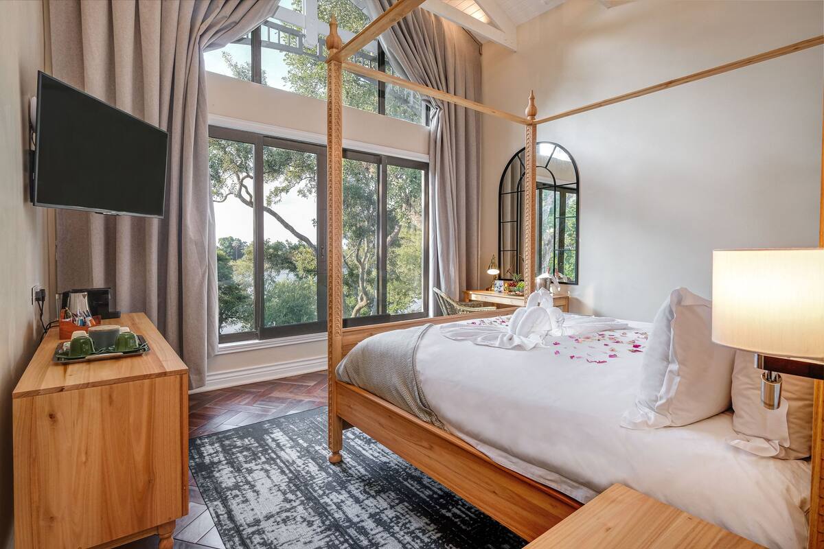 A bedroom with a four-poster bed, a flat-screen TV, a wooden nightstand, and a large window with curtains.