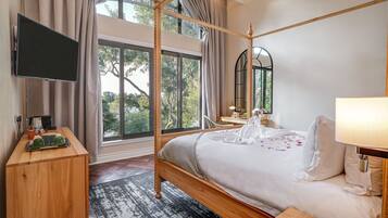 Quarto com cama de dossel, televisão de tela plana, mesa de cabeceira de madeira e uma grande janela com cortinas.