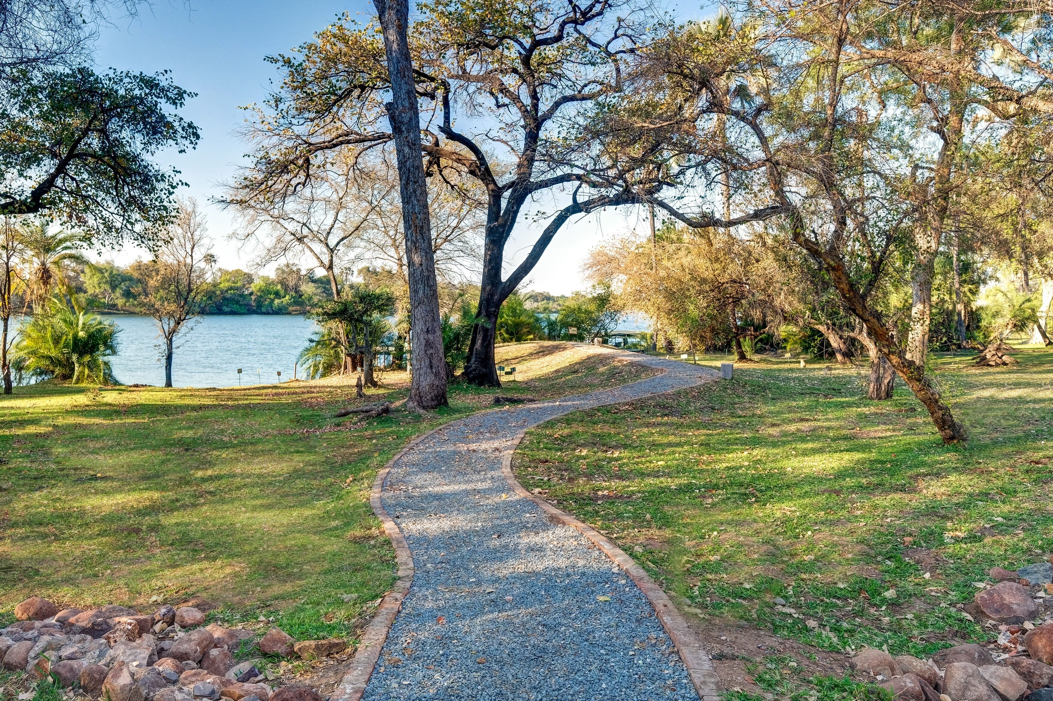 A curved gravel path in a park with trees and a lake in the background.
