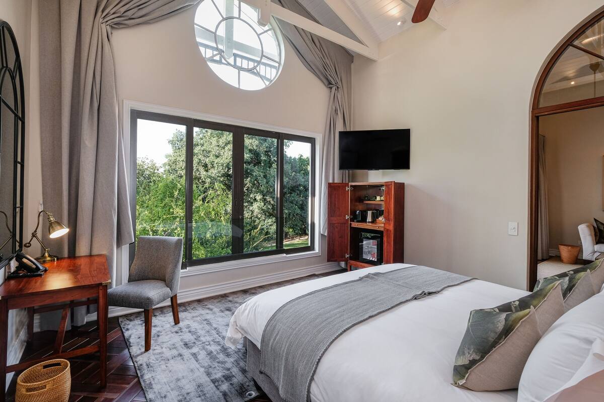 A bedroom with a large window, a bed, a desk, a chair, and a TV.