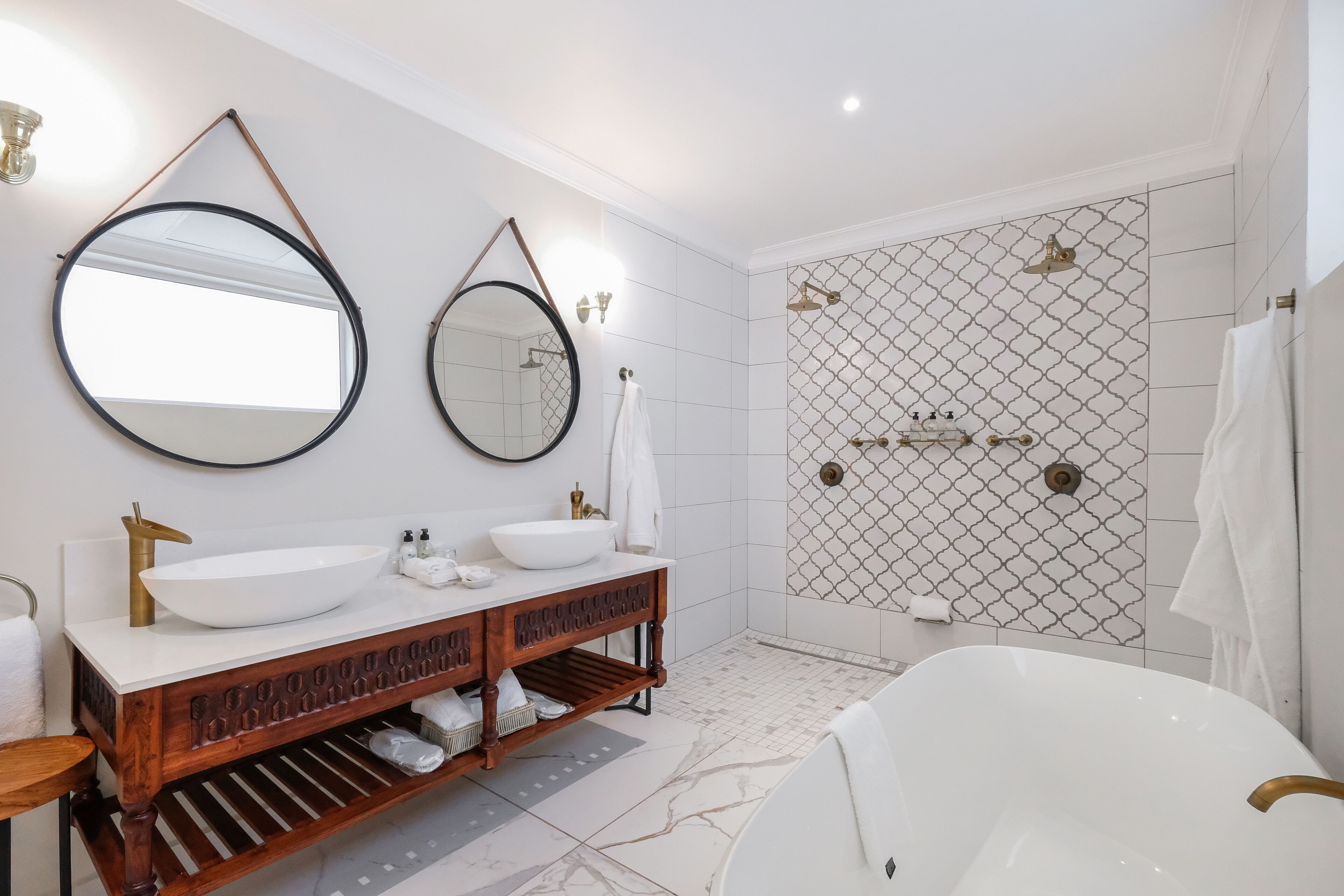 A modern bathroom with a freestanding bathtub, two sinks with round mirrors, and a shower area with a patterned tile wall.