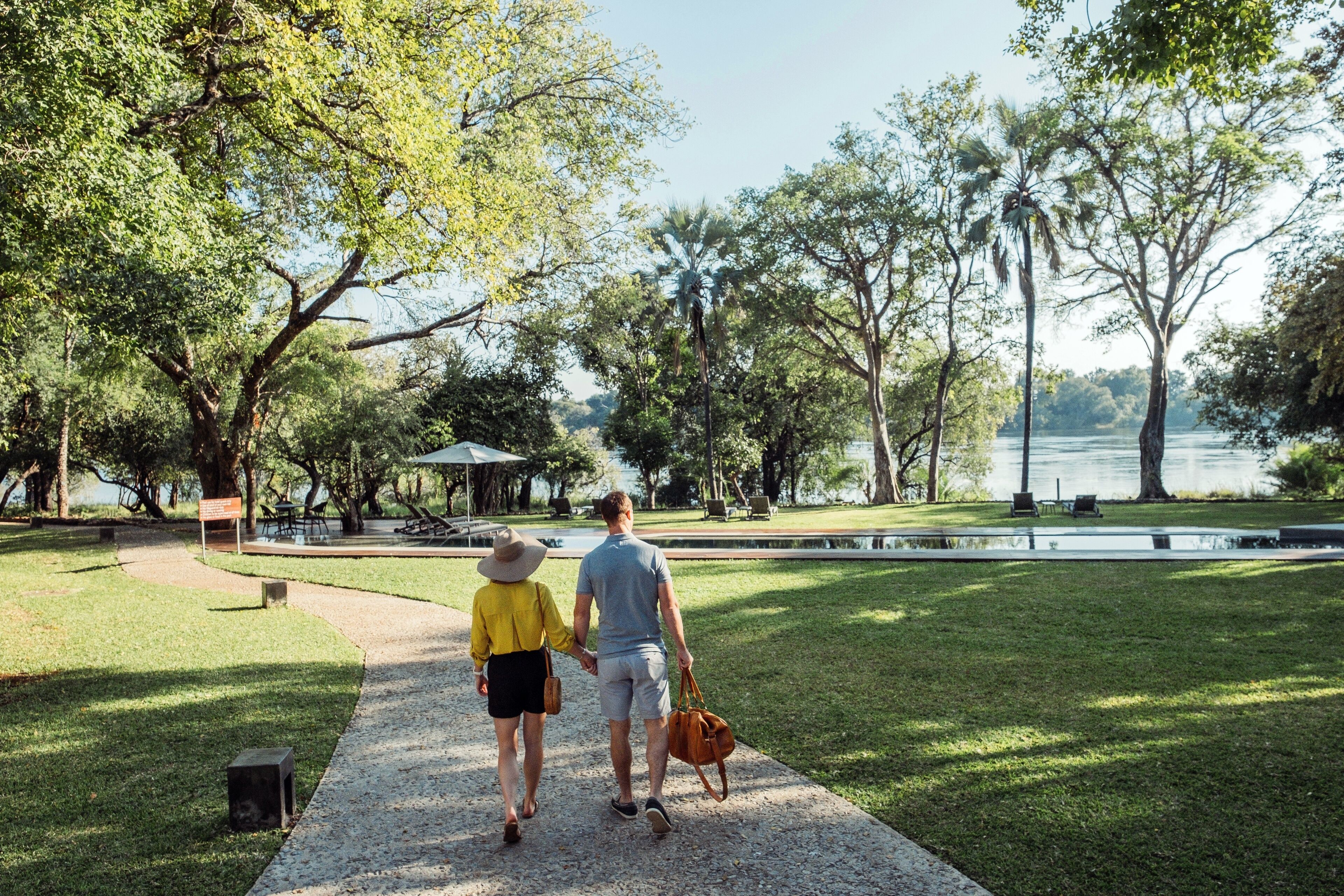 A couple walking on a paved path in a park with trees and a body of water in the background.