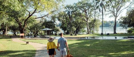 A couple walking on a paved path in a park with trees and a body of water in the background.