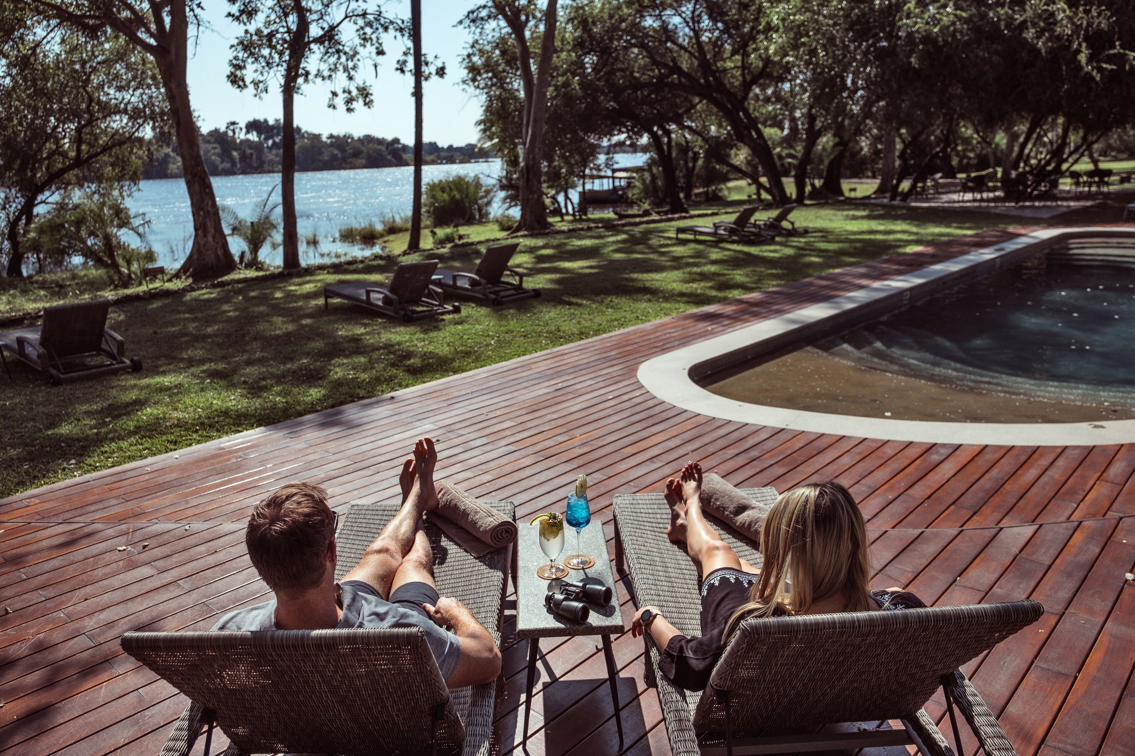 A couple relaxing on a wooden deck overlooking a lake, with lounge chairs and a table with drinks.