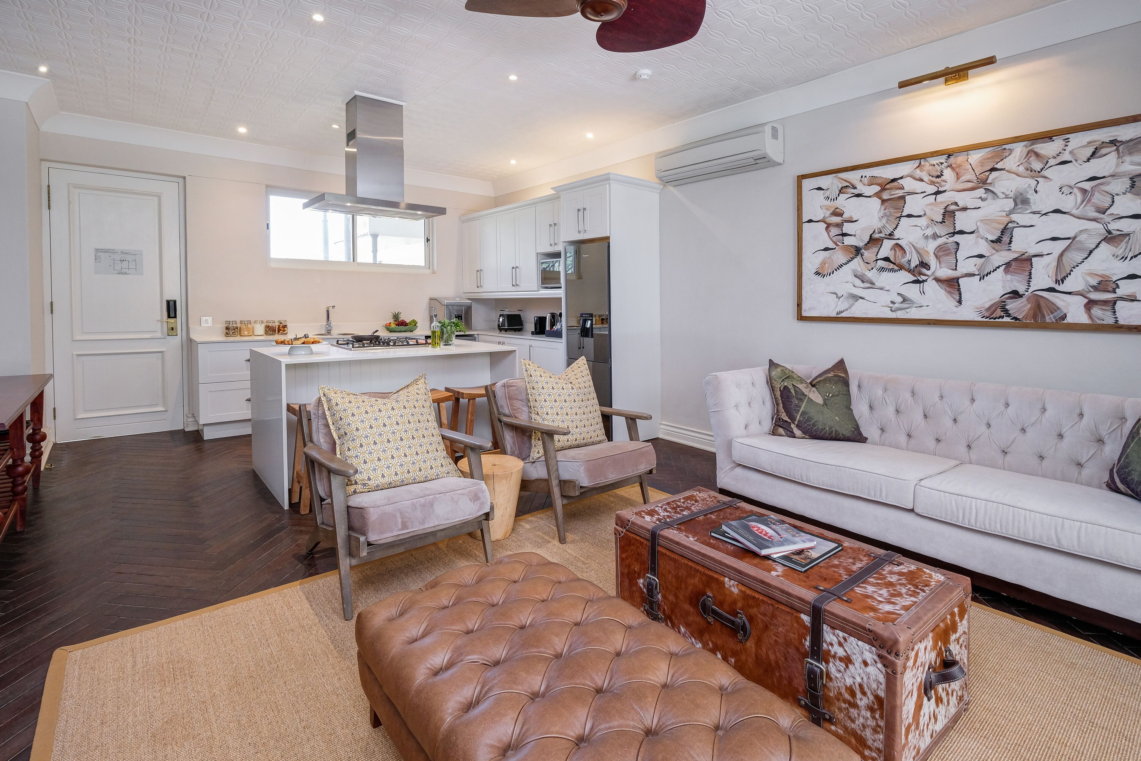 Un salon moderne avec un canapé blanc, un pouf en cuir et une table basse faite à partir d’une vieille valise. On aperçoit, au fond, une cuisine avec des meubles blancs et un coin repas avec des chaises.