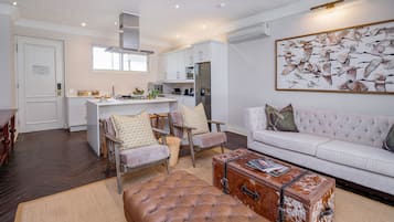 Un salon moderne avec un canapé blanc, un pouf en cuir et une table basse faite à partir d’une vieille valise. On aperçoit, au fond, une cuisine avec des meubles blancs et un coin repas avec des chaises.