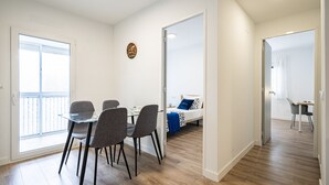 A modern room with a glass dining table, four chairs, a bed with a blue pillow, and a wooden floor.